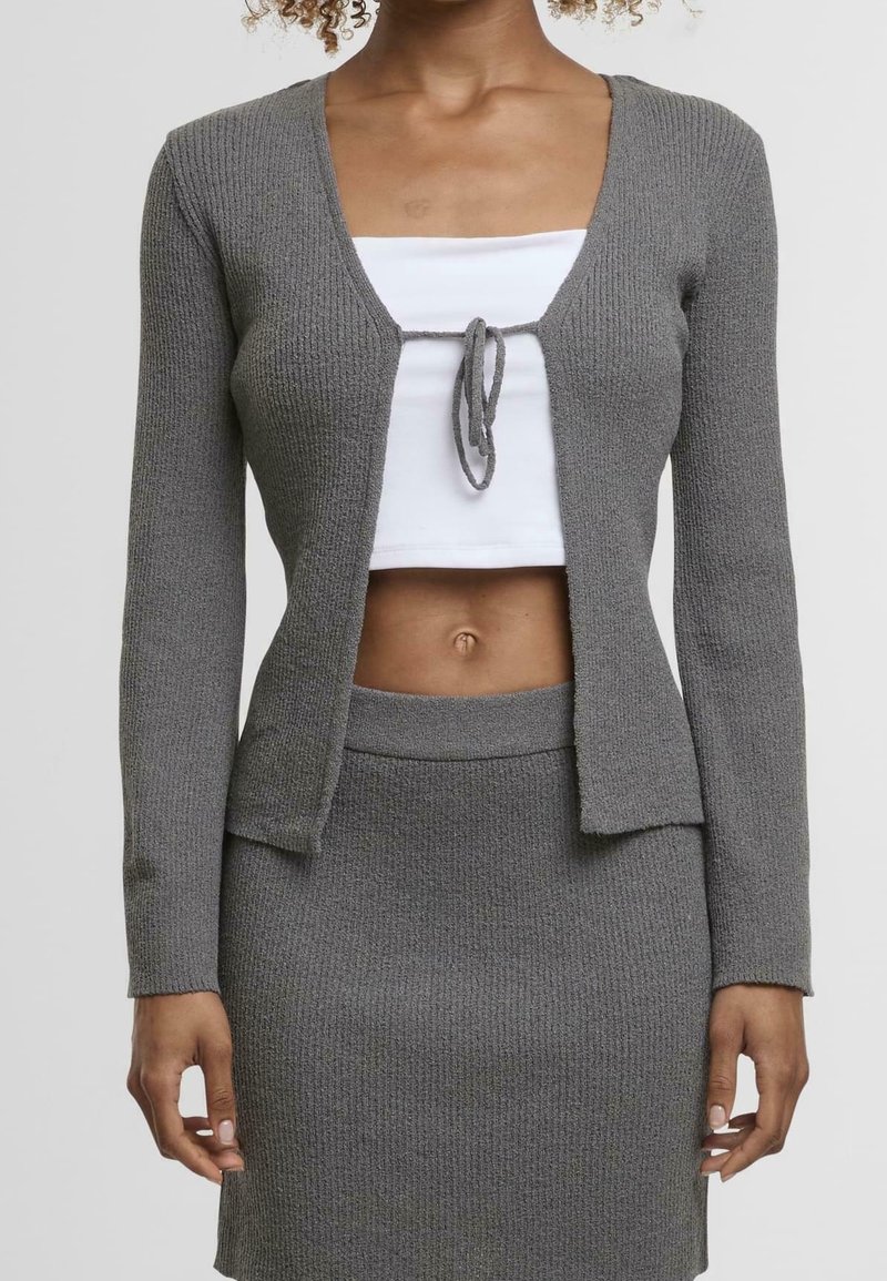 Femme portant un cardigan en maille gris noué à la poitrine, un crop top blanc et une jupe assortie en maille grise, vue du cou jusqu'à mi-cuisse.