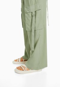 Ljusgröna vida cargo-byxor med flera fickor, tillsammans med krämfärgade sandaler med remmar som har en tjock sula och öppna tår.