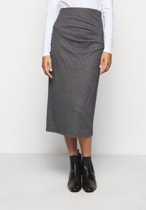 Mujer con falda midi de pata de gallo en blanco y negro, top de manga larga blanco y botines negros, de pie frente a un fondo liso.