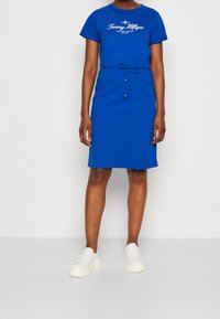 Robe t-shirt en coton bleu avec des manches courtes, impression de logo sur le devant et taille élastique avec des accents en perles. Associée avec des baskets blanches.