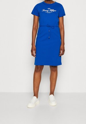 Blauwe katoenen T-shirt jurk met korte mouwen, logo-opdruk op de voorkant en een trekkoord taille met kraalaccenten. Gestyled met witte sneakers.