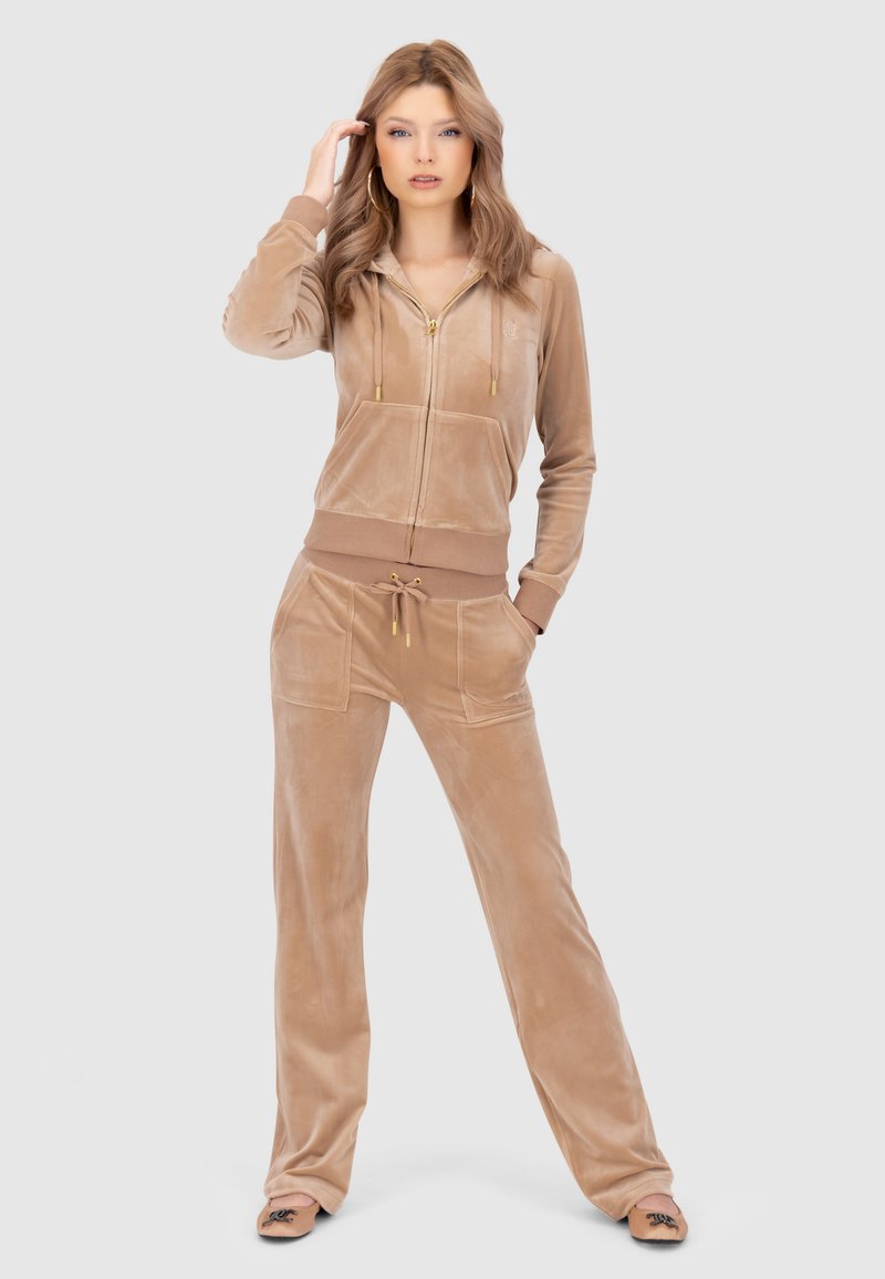 Completo tuta in velluto di colore beige chiaro con zip frontale, cappuccio e due tasche. Presenta una vita elastica e dettagli con cordoncino. Scarpe basse completano il look.