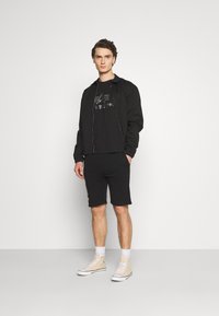 Jachetă bomber neagră peste un tricou grafic negru, asortată cu pantaloni scurți negri. Modelul poartă sneakersi bej cu glezna înaltă, pe un fundal simplu.
