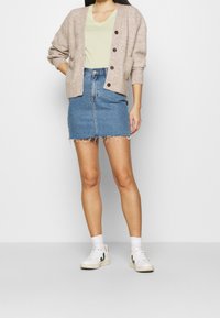 Helles beiges Strickcardigan mit braunen Knöpfen, getragen über einem hellgrünen V-Ausschnitt-Shirt, kombiniert mit einem blauen Jeans-Minirock und weißen Turnschuhen.