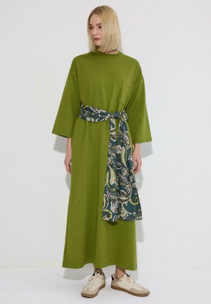 Femme aux cheveux blonds coupés au carré, portant une longue robe verte à larges manches et une ceinture à motifs, debout devant un fond blanc uni.