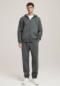 Szara bluza z zamkiem wykonaną z miękkiego materiału, w parze z dopasowanymi spodniami dresowymi. Posiada kaptur ze sznurkiem i boczne kieszenie, noszona z białymi trampkami.