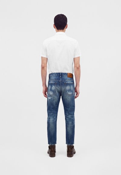 Homme debout, de dos, portant une chemise blanche à manches courtes, un jean bleu usé et des bottines marron sur un fond uni.