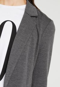 Šedý blazer s texturovanou tkaninou, s klopovým límcem a otevřeným předním dílem. V kombinaci s bílou košilí, která má velký černý grafický motiv.