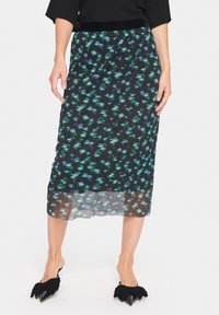 Jupe noire avec un motif floral bleu et vert, design plissé, ourlet inférieur transparent, associée à des mules noires à bout pointu avec des accents en nœud.
