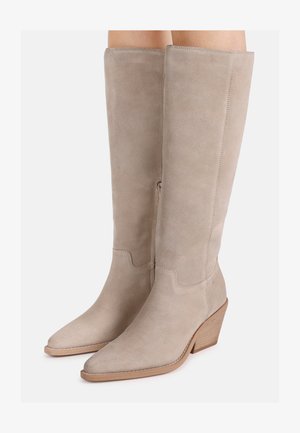 Bottes hautes en daim beige avec bouts pointus et talons carrés en bois empilés portées sur des jambes sur fond blanc.
