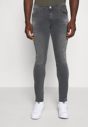 Mann in blassen grauen, eng anliegenden Jeans und weißen Adidas Sneakers, der vor einem einfarbigen, hellen Hintergrund steht.