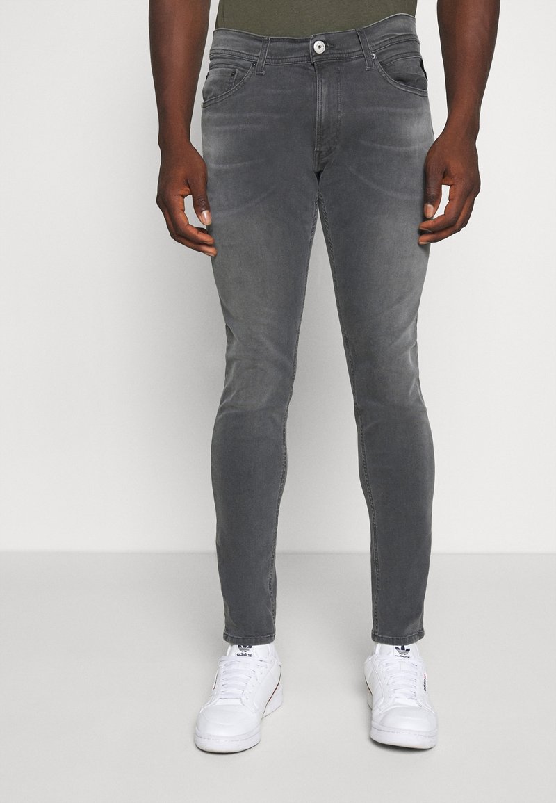 Muž v bledě šedých slim-fit džínách a bílých teniskách Adidas, stojící proti jednobarevnému světlému pozadí.