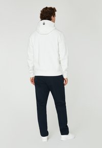Sudadera blanca con cordón y logo en la parte posterior, combinada con pantalones azul marino. El modelo lleva zapatillas blancas. Textura suave, diseño casual.