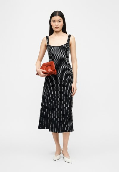 Γυναίκα που φορά ένα μαύρο μίντι φόρεμα χωρίς μανίκια με λευκές κάθετες γραμμές, κρατώντας μια κόκκινη τσάντα χειρός και φορά λευκά μυτερά παπούτσια.