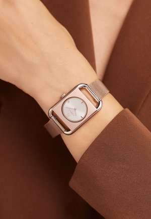 Orologio rettangolare in rosé con cinturino a rete, quadrante bianco minimalista e marker rosa. Indossato su una mano su un tessuto marrone dalla texture.