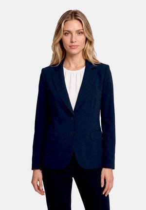 Femme aux cheveux blonds portant un blazer bleu marine foncé ajusté sur un chemisier blanc, debout devant un fond uni clair.