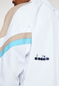 Balts apvalks ar beigtēm un zilu akcentiem, ar teksturētu audumu un izvienotu "diadora" logo uz piedurknes.
