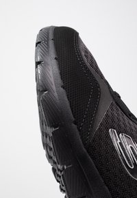 Svart atletisk sko med ett strukturerat mesh-övre, gummiyttersula och framträdande mönster. Har en slank design med reflekterande accenter.