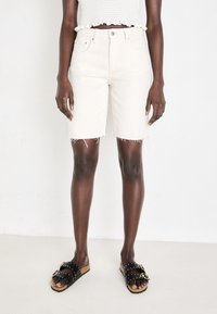 Shorts en denim couleur crème avec ourlet effiloché, taille haute, poches avant, associés à des sandales noires à semelle glissante avec des accents cloutés.