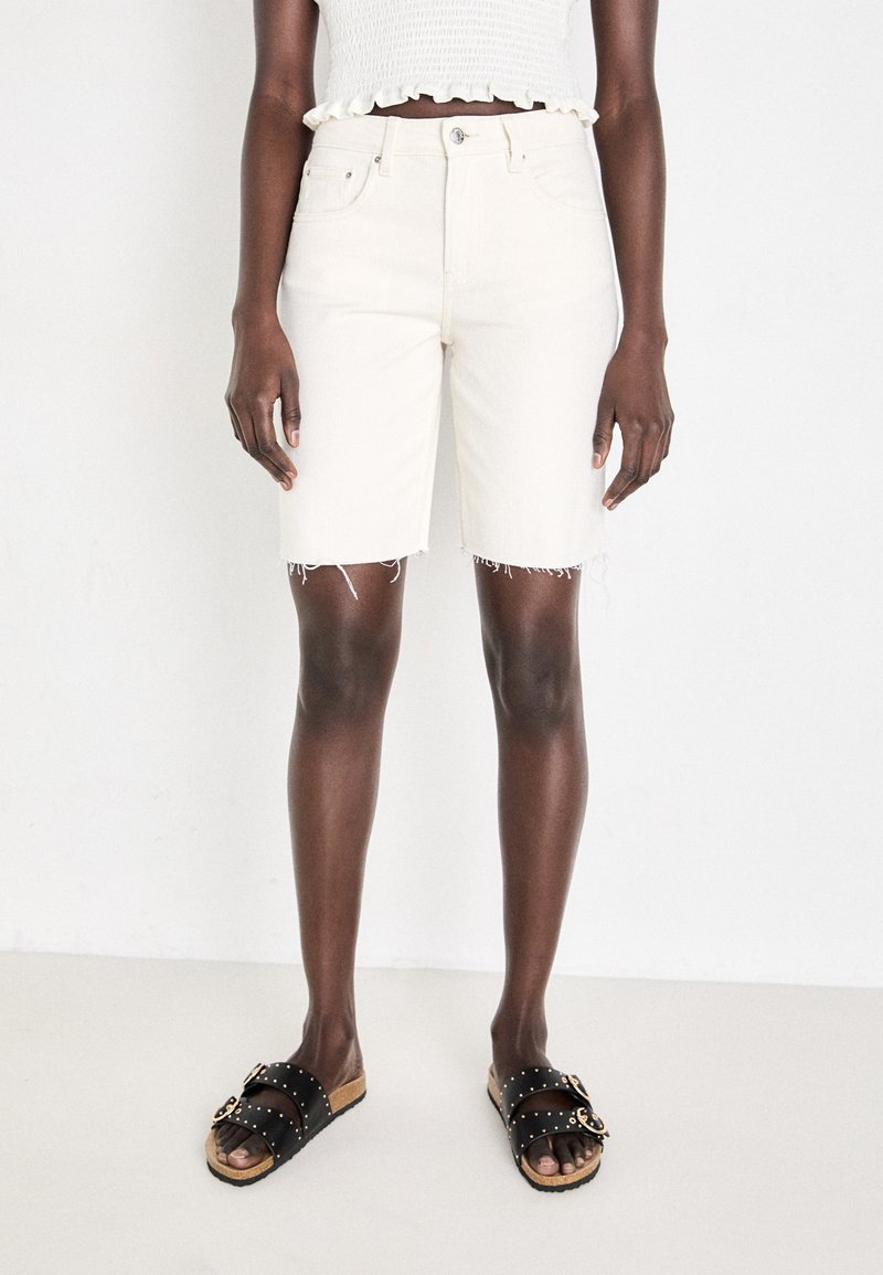 Shorts en denim couleur crème avec ourlet effiloché, taille haute, poches avant, associés à des sandales noires à semelle glissante avec des accents cloutés.