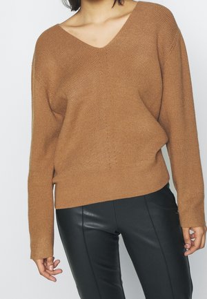 Vrouw die een bruine V-hals gebreide trui en zwarte leren broek draagt, staand tegen een effen lichte achtergrond.