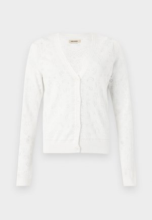 Donna che indossa occhiali da sole neri, cardigan bianco e top, gonna lunga marrone, stivali marroni e porta una grande borsa marrone a tracolla.