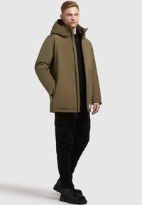 Olivgrön vadderad jacka med stor huva, dragkedjestängning, två fickor i sidan och strukturerad yta, kombinerad med svarta cargobyxor och stövlar.