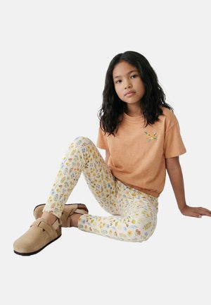 Fille aux longs cheveux foncés assise avec un bras derrière elle, portant une chemise pêche brodée de fleurs, des leggings blancs à motifs floraux et des chaussures beige sans lacets.