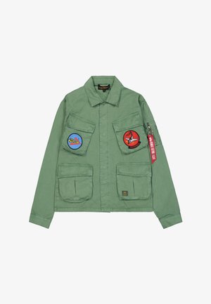 Grüne Militärjacke im Stil mit zwei großen Fronttaschen, die blaue und rote Aufnäher tragen, einem Reißverschluss in der linken Armtasche und einem Stoffetikett.
