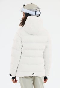 Weiße Pufferjacke mit Kapuze, ausgestattet mit einem gesteppten Design und elastischen Bündchen, getragen mit einem grauen Skihelm. Schwarzes Logodetail am Saum.