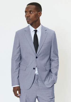 Mand iført en lysegrå jakkesæt, hvid skjorte og sort slips, med den ene hånd i lommen, kigger mod venstre mod en ensfarvet hvid baggrund.