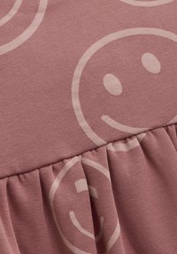 Lyserød stof med en glat tekstur, der har store beige smiley ansigt mønstre og en samlet flæsedetalje langs kanten.
