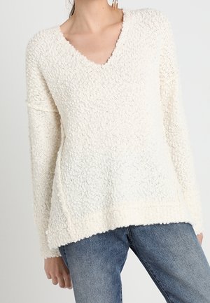 Femme portant un pull blanc texturé à col en V avec des manches longues, associé à un jean en denim bleu, debout devant un fond uni.