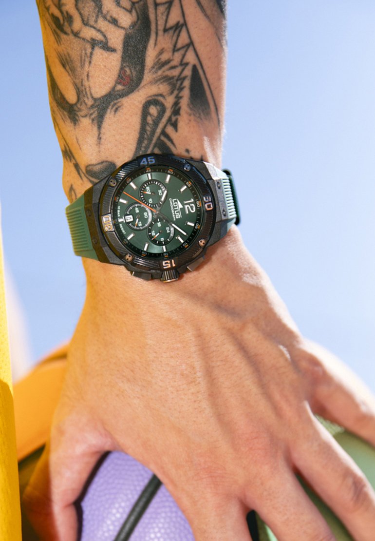 Orologio con cinturino in silicone verde, caratterizzato da una cassa nera, un quadrante rotondo, tre quadranti secondari e numeri bianchi. Una mano tiene un pallone da basket viola.