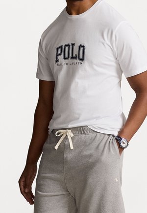 Camiseta blanca de algodón con "POLO RALPH LAUREN" impreso en azul marino. Combinada con pantalones de chándal gris claro con cordón y logo en la pierna.