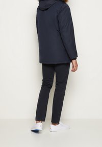 Osoba oblečená v námořnickém modrém zimním kabátu s kožešinovou kapucí, tmavých kalhotách a bílých teniskách stojící proti jednoduché světlé zdi.