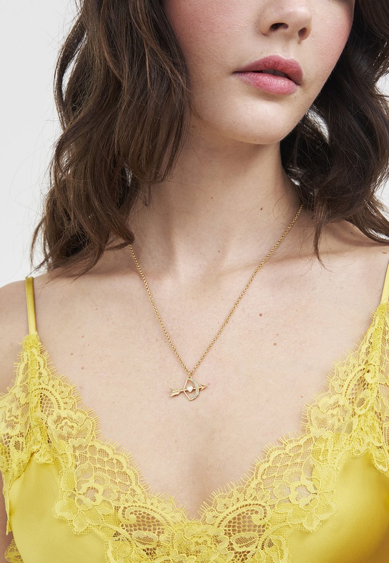Žena u žutoj majici s čipkastim rubom i zlatnom ogrlicom s privjeskom u obliku srca i strelice, prikazane vrat i dio lica te kose.