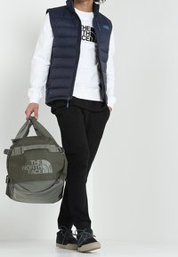 Person i svarta byxor, vit sweatshirt, marinblå väst håller en olivgrön The North Face-ryggsäck mot en vit bakgrund.