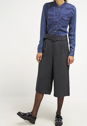 Femme portant une chemise bleue boutonnée, un pantalon gris large taille haute raccourci avec une ceinture, des collants noirs transparents et des chaussures noires brillantes.