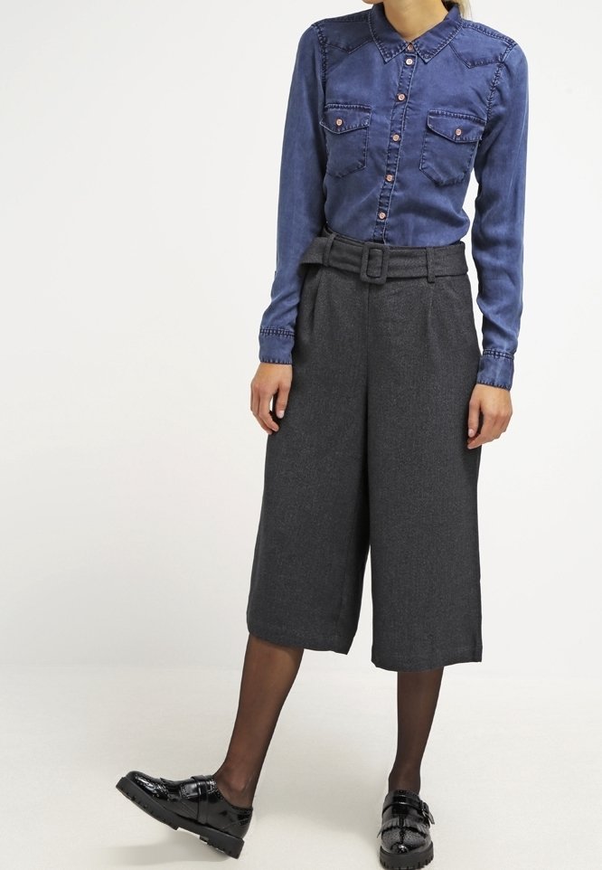 Femme portant une chemise bleue boutonnée, un pantalon gris large taille haute raccourci avec une ceinture, des collants noirs transparents et des chaussures noires brillantes.