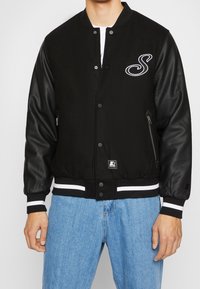 Man in een zwart varsityjack met leren mouwen, witte gestreepte manchetten, een wit "S" logo op de borst en lichtblauwe jeans.