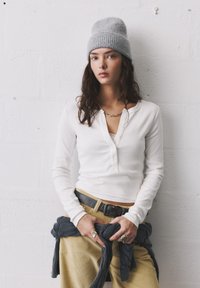 Valkoinen pitkähihainen henley-paita, jossa on napitettava kaulus, yhdistettynä keltaisiin samettihousuihin. Harmaa neulottu pipo on päässä.