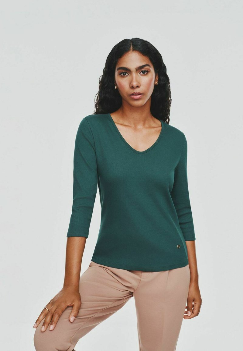 Kobieta z długimi, kręconymi włosami, ubraną w zieloną bluzkę z dekoltem w serek i 3/4 rękawami oraz beżowe spodnie, pozująca z jedną ręką na kolanie na jednolitym tle.