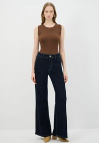 Brązowy prążkowany top na ramiączkach z ciemnoniebieskimi dżinsami typu dzwony. Dżinsy mają widoczne szwy i zapięcie na guzik z przodu. Modelka nosi beżowe szpilki.