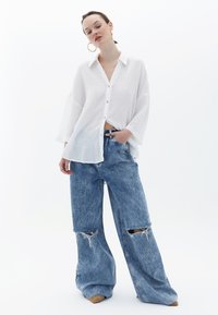Camicia bianca in cotone, ampia e con bottoni, dotata di colletto; abbinata a jeans in denim blu larghi e strappati con buchi alle ginocchia.