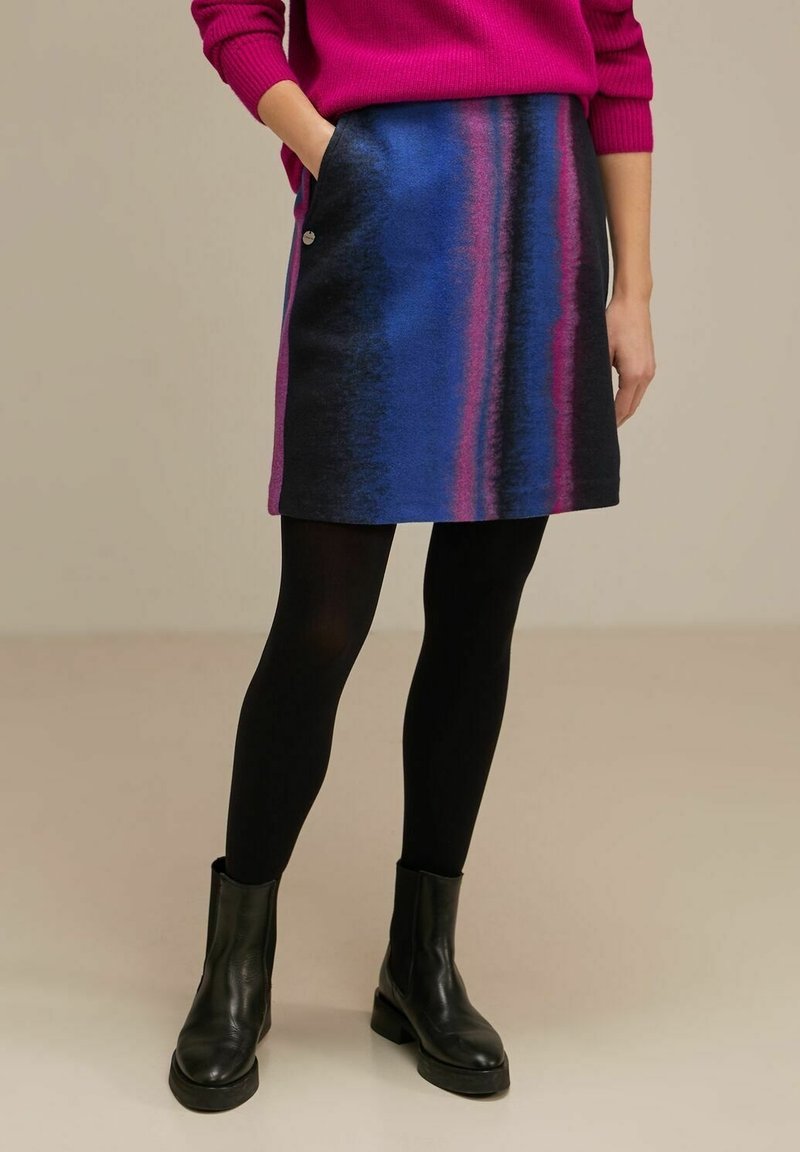 Une jupe A-line multicolore avec des rayures bleues, noires et roses, dotée de poches latérales et d'un accent de bouton métallique. Portée avec des collants noirs et des bottes.