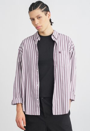 Līdzceļš, svītrains krekls lavandas un baltā krāsā ar pogu priekšā un kabatu, uzvilkts virs melna T-krekla. Brīvs piegriezums.