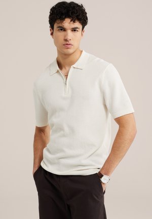 Noor mees lokkis juustega, kandes valget lühikeste varrukatega tekstuurilise polo särki ja tumedaid pükse, käed taskutes, neutraalne taust.