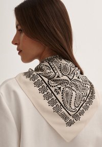 Kremowy szalik z czarnym wzorem paisley i ozdobnymi brzegami, zarzucony na szyję i ramiona. Gładka faktura materiału.