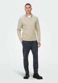 Jersey de punto acanalado beige con cuello de medio cremallera, combinado con pantalones de cuadros oscuros y botas negras de tobillo. Paleta de colores neutros.